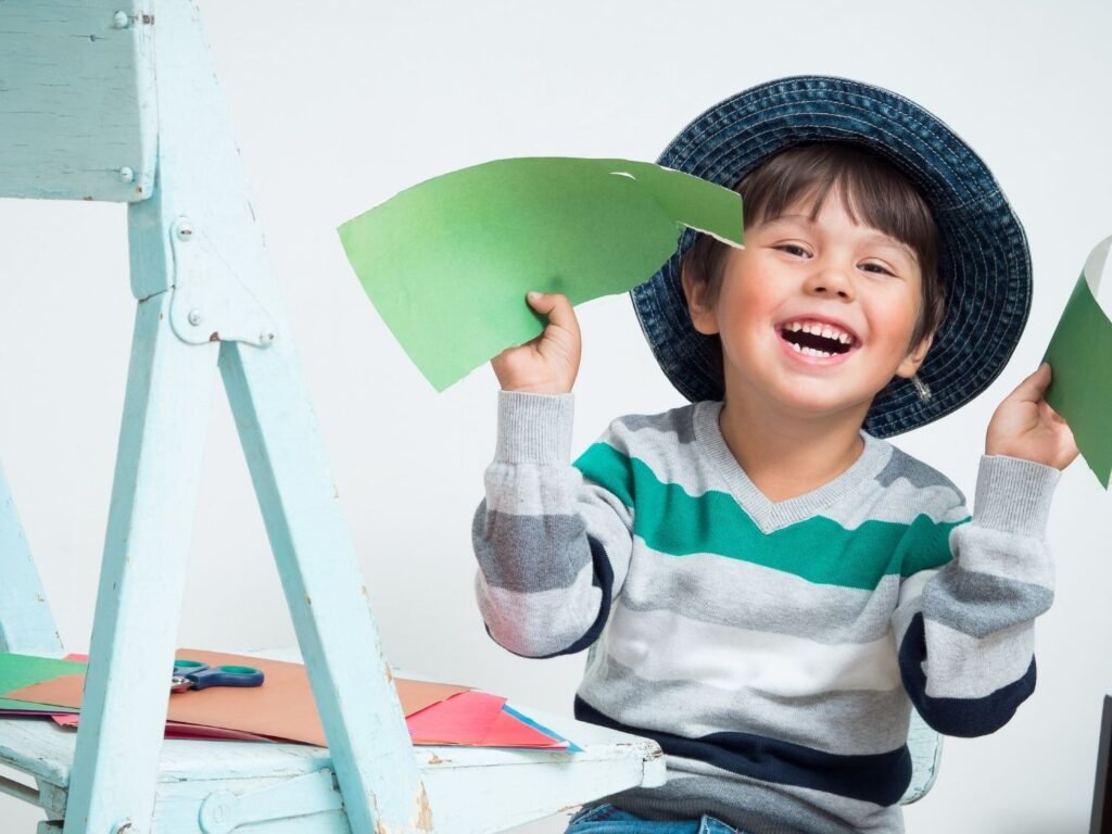 Petit garçon souriant tenant des morceaux de papier vert découpés lors d'une activité de motricité fine.