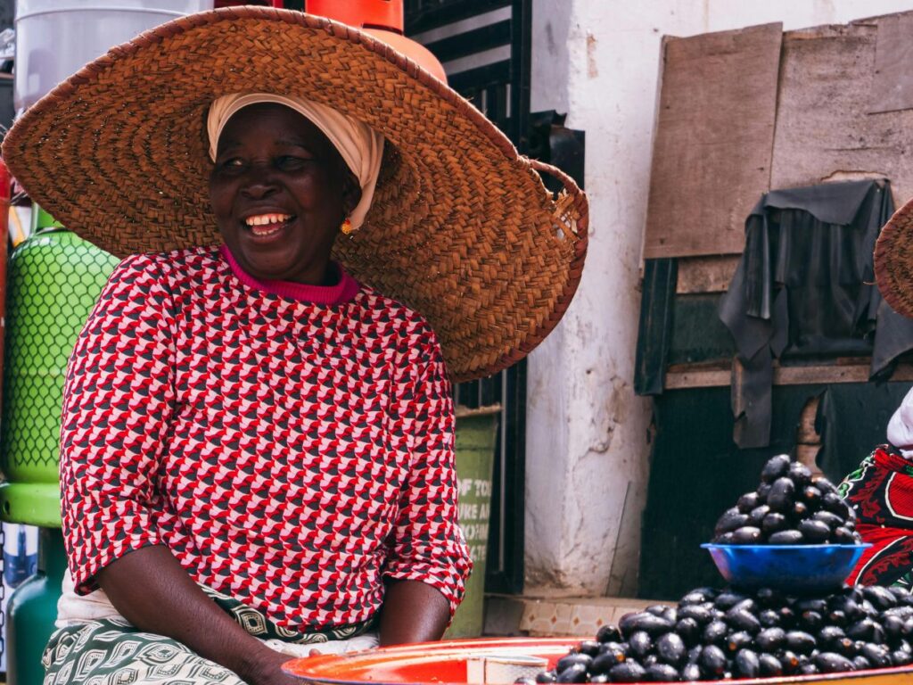 Une marchande africaine souriante portant un grand chapeau de paille traditionnel sur un marché local.