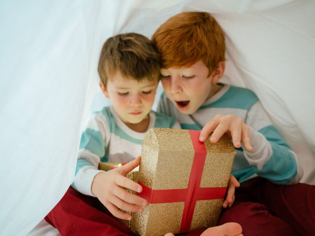 Deux jeunes enfants sous une cabane en draps blancs, l'un d'eux exprime une vive émotion de surprise en ouvrant un cadeau lumineux.