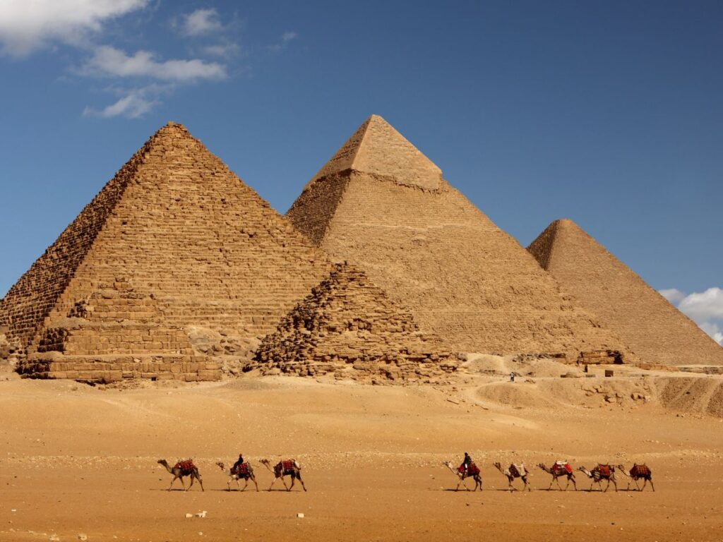 Alignement des trois grandes pyramides de Gizeh avec une caravane de dromadaires marchant dans le désert sous un ciel bleu.