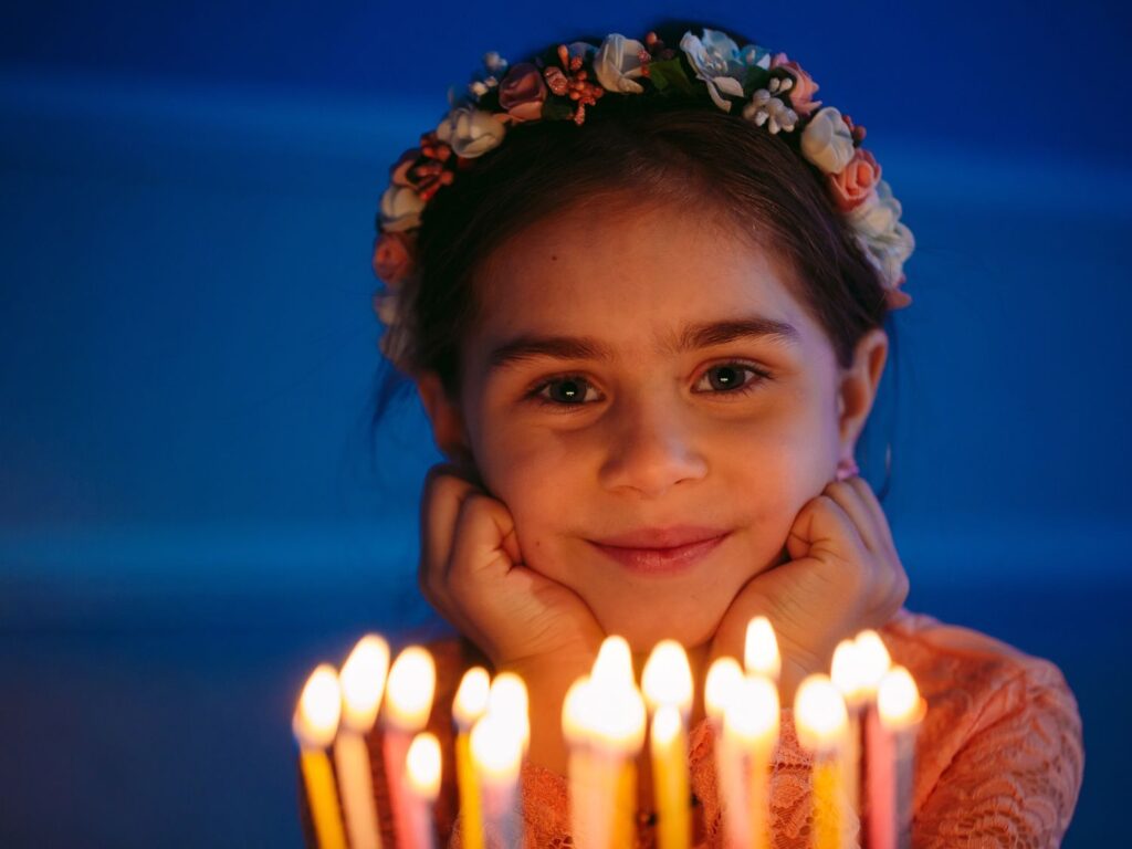 Petite fille souriante portant une couronne de fleurs, le visage illuminé par la joie devant les bougies allumées de son gâteau d'anniversaire.