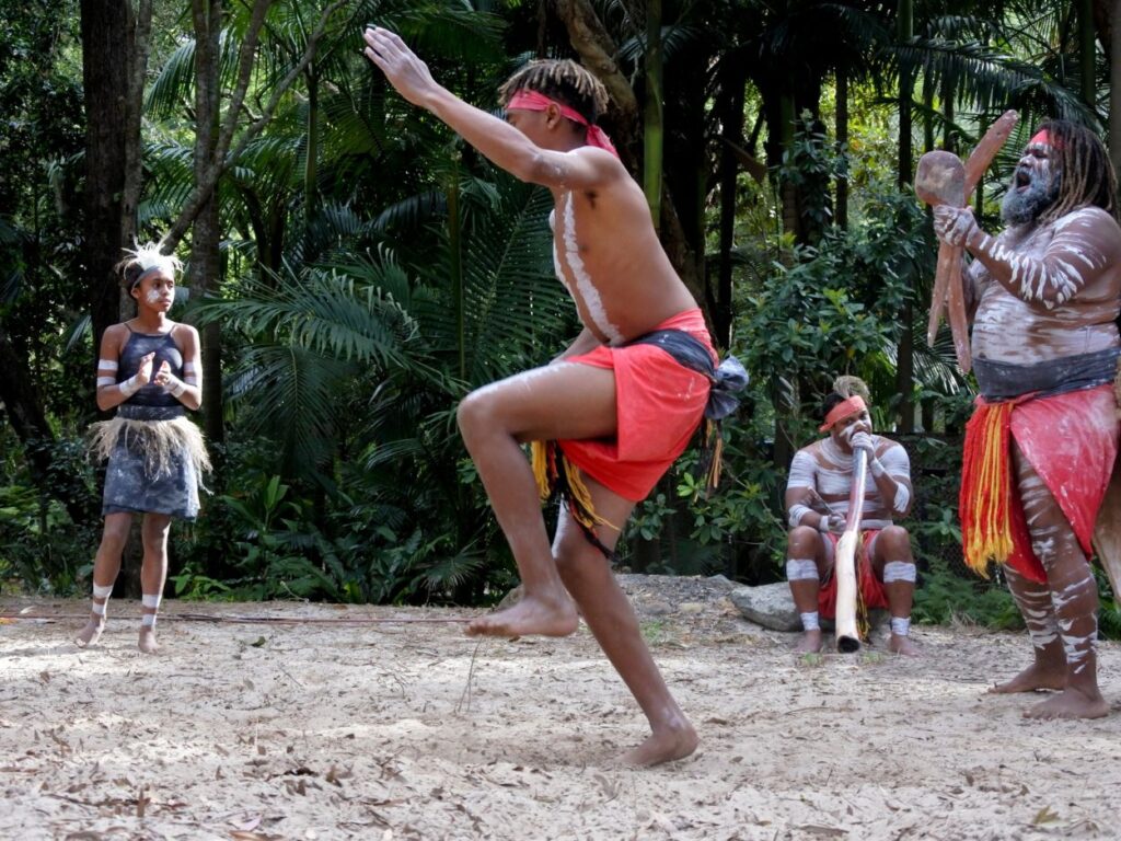 Groupe d'Aborigènes en tenue traditionnelle pratiquant une danse et jouant du didgeridoo en pleine nature.