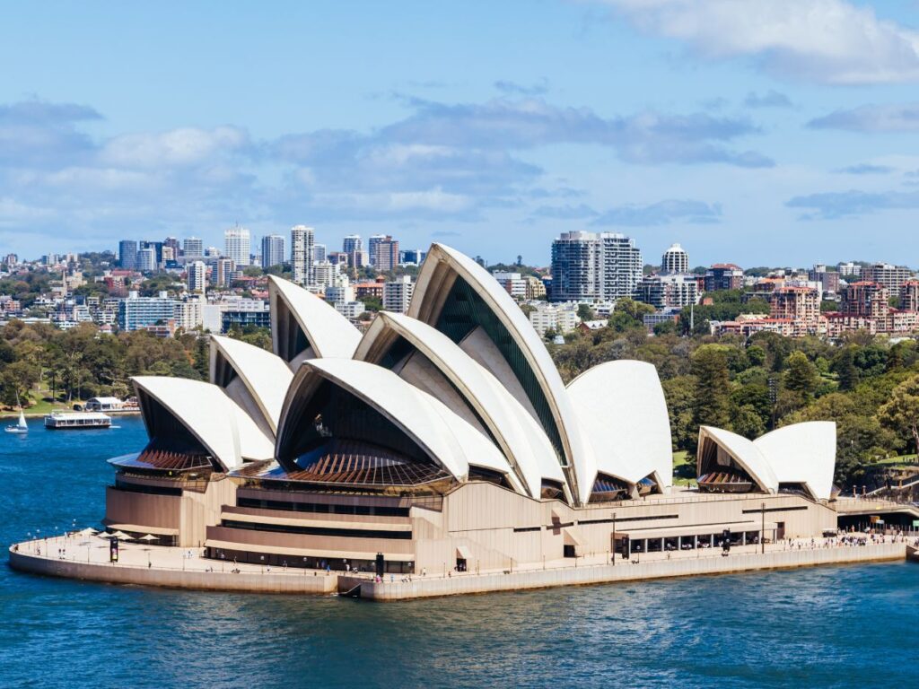 Vue panoramique de l'Opéra de Sydney avec ses toits blancs caractéristiques surplombant le port, sur fond de gratte-ciel.